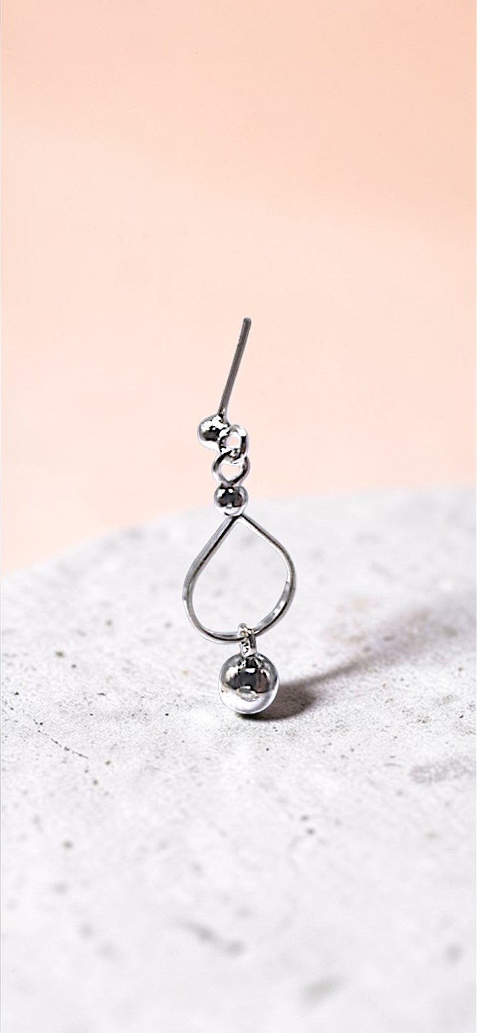Silver teardrop-shaped pendant with a small round charm on a textured surface with a pink gradient background.